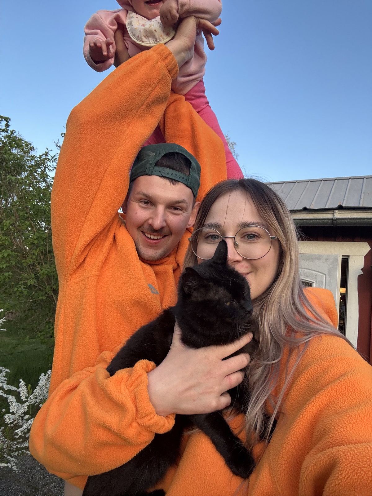 Two people in orange jackets holding a baby and a black cat outdoors.